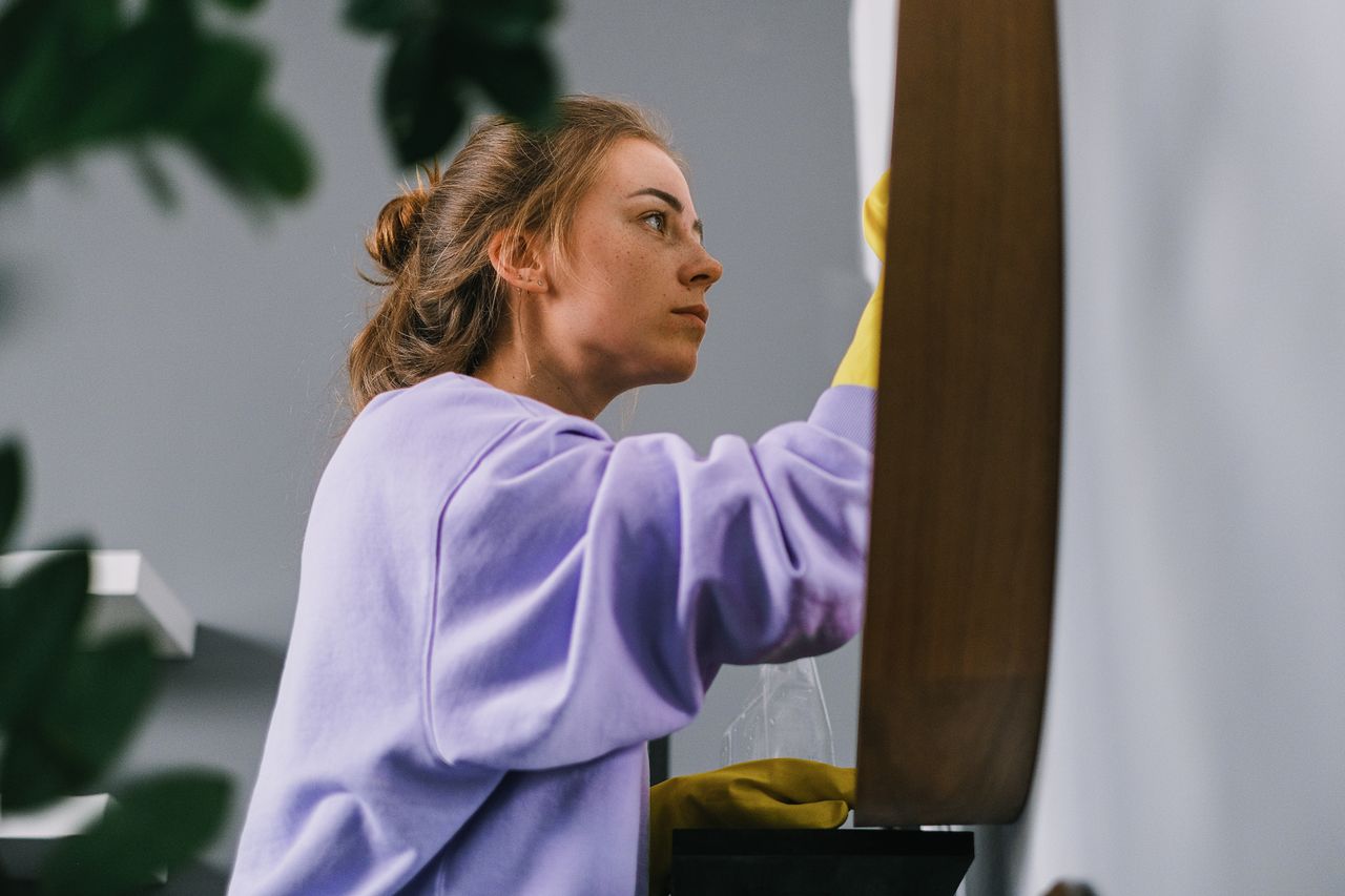 Vue de côté d’une jeune femme concentrée, en gants en latex, debout et nettoyant un miroir dans une pièce lumineuse en journée